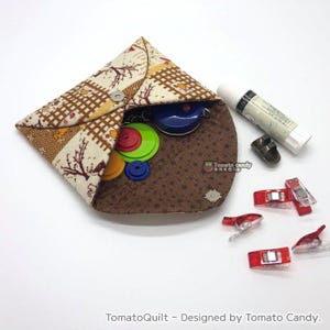 May include: A brown and beige fabric pouch with a snap closure, open to reveal a variety of sewing supplies, including buttons, a measuring tape, and a glue stick. The pouch is decorated with a plaid pattern and a floral design. The text "TomatoQuilt - Designed by Tomato Candy." is visible at the bottom of the image.