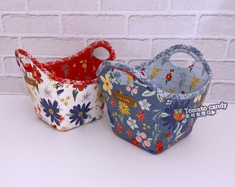 May include: Two quilted fabric baskets with cut-out handles. One basket features a white exterior with red and blue floral patterns and a red interior. The other is blue with multi-colored flowers and a light blue interior. Both have Tomato candy labels.