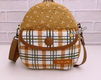Puede incluir: Una mochila acolchada de color amarillo mostaza con un bolsillo frontal de cuadros blancos, verdes y marrones. El bolso cuenta con un cierre de botón marrón, una correa marrón y una etiqueta rectangular con el texto Tomato candy. Se ve un pequeño logo con el texto Tomato candy.