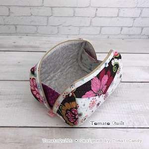 May include: A quilted fabric pouch with a gold-tone zipper, featuring a vibrant floral pattern in shades of pink, yellow, and black on a white background. The interior is lined with a light gray fabric with a subtle white leaf print pattern.