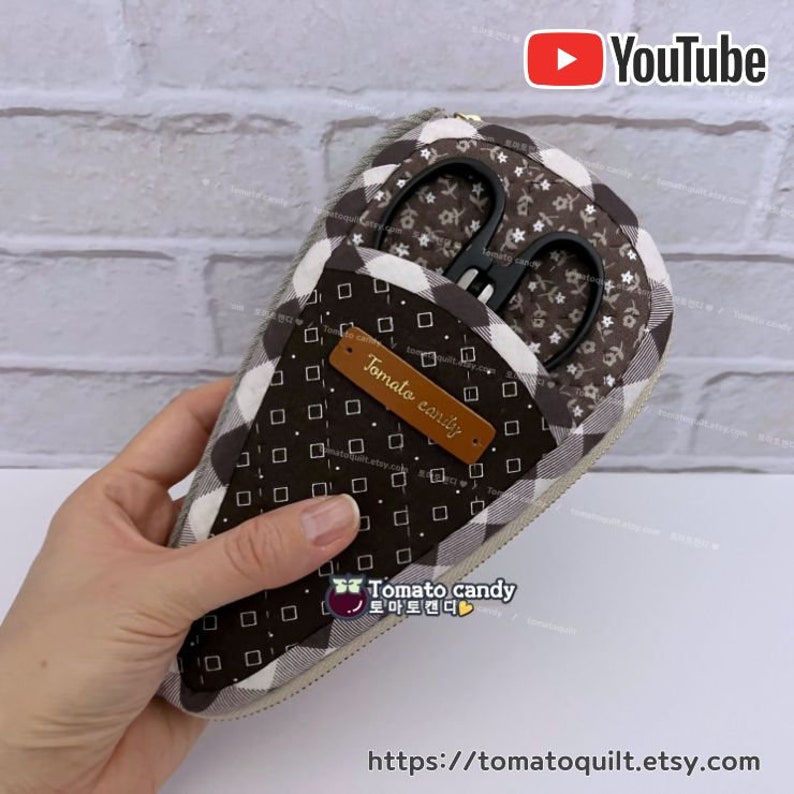 May include: A brown and white checkered fabric scissor case with a zipper closure. The case has a small pocket on the inside for storing small items. The case is labeled "Tomato candy" and has a website address on the bottom.