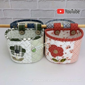 May include: Two quilted fabric hanging baskets with floral designs. The basket on the left has a green and white floral design with a black and white plaid flower. The basket on the right has a red and white floral design with a red flower. Both baskets have a wooden dowel rod at the top and two button closures.