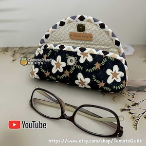 Può includere: Un astuccio per occhiali in tessuto semicircolare con sfondo nero, motivi a fiori e stelle bianchi e beige, un bordo a chevron e una chiusura a scatto in metallo. Un'etichetta in pelle marrone riporta la scritta Tomato candy. Occhiali marroni.