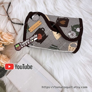 May include: A gray fabric pouch with a brown trim and a black and white checkered pattern. The pouch has a brown label that says "Tomato candy" and a brown sticker with a tomato and the text "Tomato candy" in Korean. The pouch is on a white fuzzy surface.