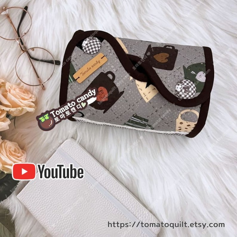 May include: A gray fabric pouch with a brown trim and a black and white checkered pattern. The pouch has a brown label that says "Tomato candy" and a brown sticker with a tomato and the text "Tomato candy" in Korean. The pouch is on a white fuzzy surface.