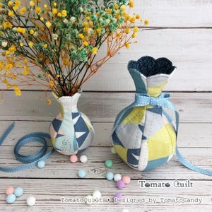 May include: Two fabric vases with a quilted geometric pattern in shades of blue, yellow, and white. The larger vase features a light blue ribbon tied around its neck. The text Tomato Quilt Designed by TomatoCandy is visible at the bottom.
