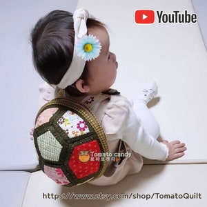 May include: A baby wearing a white shirt and a headband with a flower on it. The baby is wearing a patchwork quilt backpack in the shape of a turtle. The backpack is made of different fabrics and colors, including green, red, pink, and white. The backpack has a small, orange, cartoon tomato with the text "Tomato candy" in Korean on it. The text "https://www.etsy.com/shop/TomatoQuilt" is at the bottom of the image.