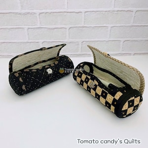May include: Two quilted fabric pencil cases with different patterns. One case is black with gold stars and the other is black and white checkered with gold stars. Both cases are open and have a white background.