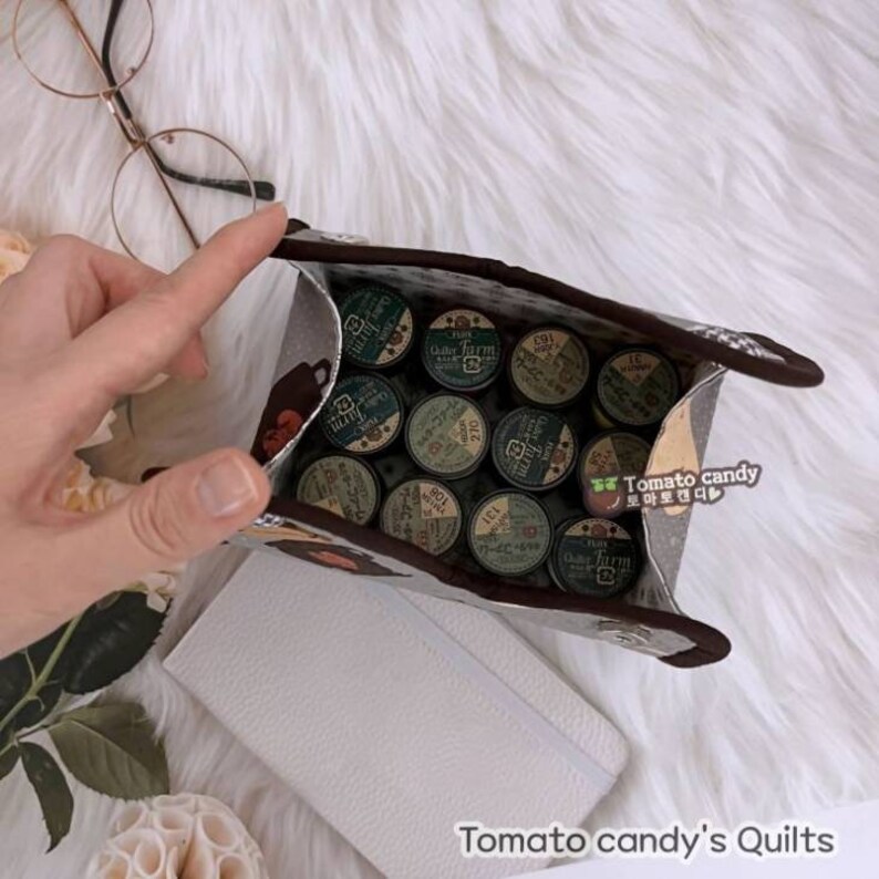 May include: A gray and brown fabric pouch with a floral pattern and a bear design. The pouch is open and filled with small, round, green and white spools of thread. The text "Gallo Farm" is visible on the spools.