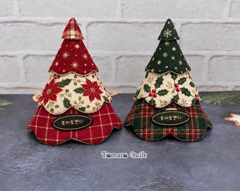 Könnte beinhalten: Zwei weihnachtliche Baumschmuck-Objekte aus Stoff mit gewelltem Rand. Der linke Baum zeigt rote Karomuster, Weihnachtssterne und goldene Sterne. Der rechte Baum zeigt grüne Karomuster, Stechpalmen und Schneeflocken. Beide haben schwarze Etiketten.