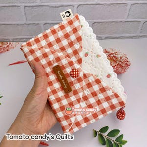 Può includere: Una busta di tessuto gingham rosso e bianco con un'etichetta marrone che dice "Tomato candy" e un bottone rosso. La busta ha un bordo di pizzo bianco e un fiocco rosso.