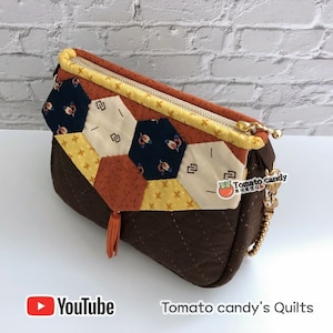 Może przedstawiać: Brązowa pikowana torba z tkaniny z zamkiem błyskawicznym i łańcuszkowym paskiem. Torba ma patchworkowy wzór z pomarańczowej, żółtej i niebieskiej tkaniny. Torba ma frędzle na dole i mały brelok z pomidorem. Brelok ma napis "Tomato candy". Torba leży na białym tle z ceglaną ścianą w tle. Pod torbą znajduje się napis "Tomato candy's Quilts".