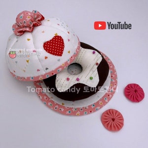 May include: A white and pink fabric donut pincushion with a red heart and a brown fabric donut base. The pincushion is decorated with small white hearts and a star. The base is decorated with a star and small white dots. The pincushion is surrounded by two small pink fabric flowers.