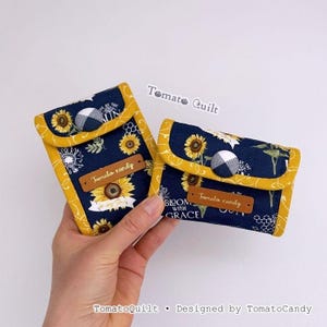 May include: Two small, navy blue fabric pouches with yellow trim and sunflower patterns. Each pouch has a button closure and a small, brown label that reads "Tomato candy". The pouches are held in a hand.