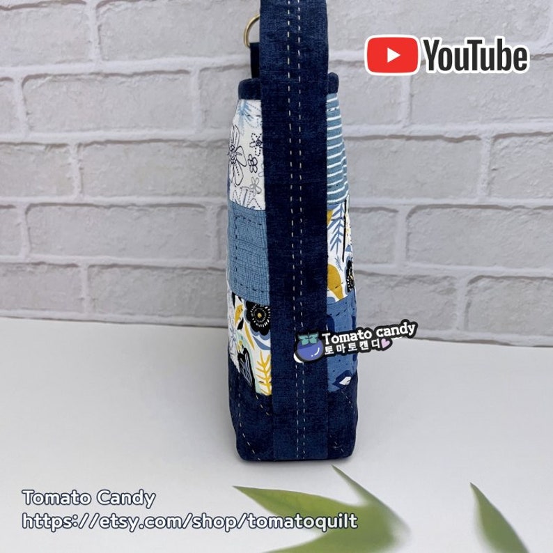 Pode incluir: Uma bolsa tote de tecido patchwork azul e branco com uma al&ccedil;a de brim. A bolsa tem um padr&atilde;o floral e um pequeno bolso na frente. A bolsa est&aacute; sobre uma superf&iacute;cie branca com uma folha verde.