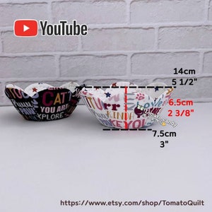 Peut inclure: Deux bols en tissu avec un bord en forme de fleur. Le bol de gauche est noir avec du texte blanc et le bol de droite est blanc avec du texte color&eacute;. Les bols mesurent 14 cm de large et 7,5 cm de haut.
