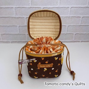 Może przedstawiać: Brązowa i pomarańczowa torba ze sznurkiem do ściągania z podszewką w kratkę. Torba ma zamek błyskawiczny i etykietę z napisem "Tomato candy".