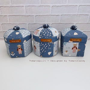 Può includere: Tre scatole di stoffa fatte a mano con disegni patchwork blu e bianchi. Ogni scatola presenta un coperchio a cupola con un pomello blu scuro e un'etichetta in pelle con la scritta "Tomato candy". Le scatole sono decorate con motivi di orsi e fiori.