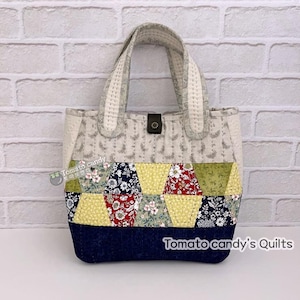 Op de afbeelding: Een patchwork draagtas met een blauwe denim bodem en een lichtgrijze stof met een bloemenpatroon. De tas heeft twee handvatten en een kleine bruine knoopsluiting. De tas is gelabeld met "Tomato candy's Quilts".