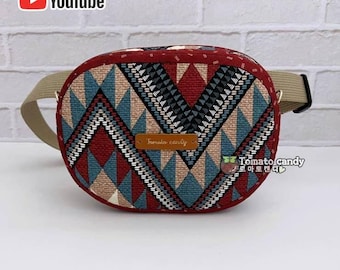 Puede incluir: Una riñonera de forma ovalada con un patrón geométrico en tonos rojo, azul y beige. Una etiqueta rectangular marrón con el texto Tomato Candy está centrada en la parte frontal. El bolso incluye una correa beige ajustable y una hebilla.