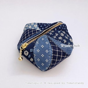 Puede incluir: Una pequeña bolsa acolchada en forma de estrella hecha de telas en tonos azules con patrones blancos, incluyendo flores, puntos y cuadros. Cuenta con una cremallera metálica dorada y el texto TomatoQuilt.