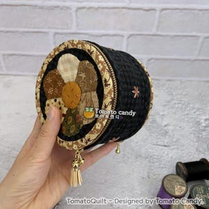 Puede incluir: Un estuche redondo de tela negra con cremallera, que presenta un diseño de flor acolchada en la tapa con pétalos de tela estampada en beige, marrón y verde. El lateral es negro con una pequeña flor bordada y un tirador de cremallera dorado.