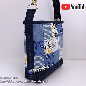 Pode incluir: Uma bolsa tote de tecido patchwork azul e branco com uma al&ccedil;a azul marinho e ferragens douradas. A bolsa tem um padr&atilde;o floral e geom&eacute;trico. O texto "Tomato Candy" e um endere&ccedil;o de site est&atilde;o impressos na parte inferior da bolsa.