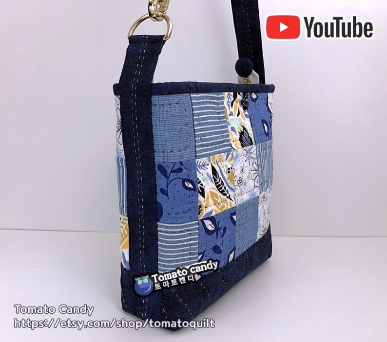 Pode incluir: Uma bolsa tote de tecido patchwork azul e branco com uma al&ccedil;a azul marinho e ferragens douradas. A bolsa tem um padr&atilde;o floral e geom&eacute;trico. O texto "Tomato Candy" e um endere&ccedil;o de site est&atilde;o impressos na parte inferior da bolsa.