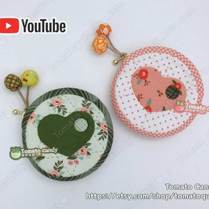 K&ouml;nnte beinhalten: Zwei runde M&uuml;nzbeutel mit herzf&ouml;rmigen Stoffakzenten. Ein Beutel ist gr&uuml;n mit einem wei&szlig;en Blumenmuster und der andere ist rosa mit wei&szlig;en Punkten. Beide Beutel haben einen kleinen Knopf und eine Stoffschlaufe mit einem kleinen orangenen Pompon.