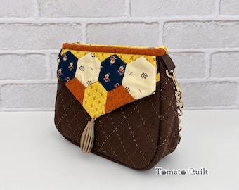 Puede incluir: Un bolso bandolera acolchado de color marrón con una solapa de diseño de patchwork hexagonal en amarillo, crema y azul marino. El bolso incluye una correa de cadena dorada, un tirador de borla beige y el texto TomatoQuilt Designed by TomatoCandy.