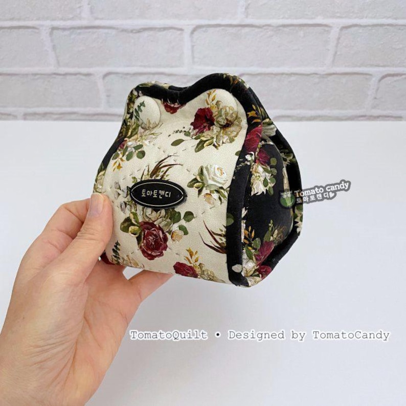 May include: A floral print pouch with a black trim, held in a hand. The pouch features a cream background with red, white, and green floral patterns. The text "TomatoCandy" is visible on the pouch.