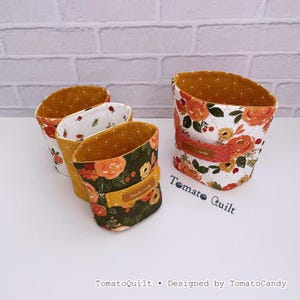 May include: Three fabric storage baskets with quilted textures. One features a white background with orange floral patterns, while the others have dark green and mustard yellow floral prints. Each basket includes a small rectangular label reading Tomato Candy.
