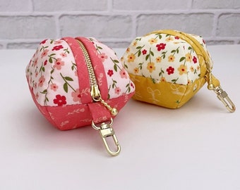 Puede incluir: Dos pequeños estuches de tela con cremalleras doradas y mosquetones de metal. Uno tiene un estampado floral rosa con detalles en rosa sólido, mientras que el otro tiene un estampado floral amarillo con detalles en amarillo sólido. El texto dice Tomato candy's Quilts.