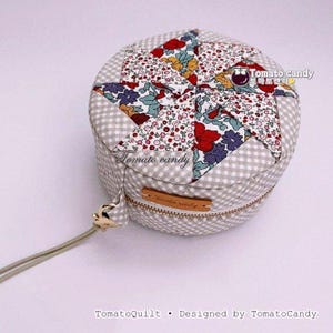 Könnte beinhalten: Ein rundes Stoffetui mit Reißverschluss, das an den Seiten ein grau-weißes Vichy-Muster und auf der Oberseite ein mehrfarbiges florales Patchwork-Sternmuster aufweist. Ein kleines braunes Lederetikett mit dem Text Tomato candy ist vorne angebracht.
