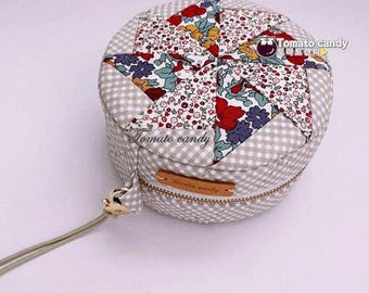 Puede incluir: Un estuche redondo de tela con cremallera que presenta un patrón de cuadros vichy gris y blanco en los laterales y un diseño de estrella de patchwork floral multicolor en la parte superior. Una pequeña etiqueta de cuero marrón con el texto Tomato candy.