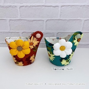 May include: Two fabric baskets with floral patterns, one in rust orange and one in teal green. Each basket features a large, dimensional fabric flower attached to the front and a small button accent on the side. The text TomatoQuilt is visible.