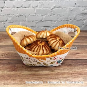 Puede incluir: Una cesta de tela con base blanca y un patrón multicolor de frutas y hojas, acentuada por un borde naranja con estampado de estrellas blancas. La cesta contiene varias galletas estriadas de color marrón dorado. El texto TomatoQuilt es visible.