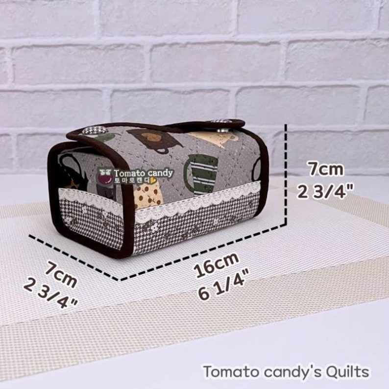 May include: A gray and brown fabric tissue box cover with a white lace trim. The cover features a pattern of teacups and bears. The cover measures 7 inches by 6 1/4 inches.