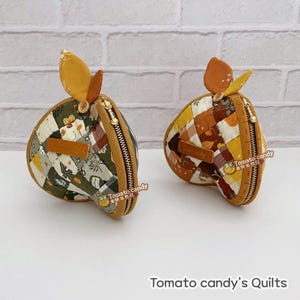 May include: Two quilted fabric pouches shaped like apples, featuring a patchwork design in shades of green, brown, yellow, and white. Each pouch has a gold zip, a small brown leaf accent, and a rectangular leather label reading Tomato candy.