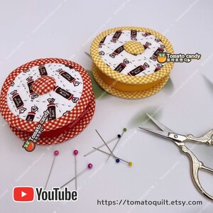 May include: Two fabric pincushions with a red and white checkered pattern and a yellow and white checkered pattern. The pincushions are shaped like spools of thread and have a button on top. The pincushions are surrounded by sewing pins and a pair of gold scissors. The text "Tomato candy" is printed on the pincushions.