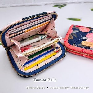 May include: Two zippered wallets with a dark blue exterior featuring a pink floral pattern and a light pink interior with a floral print. The open wallet displays multiple card slots, a central zippered coin pocket, and a gold-tone zipper.