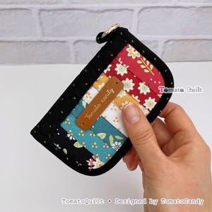 May include: A small, rectangular fabric pouch with a black border and a multi-coloured quilted front featuring floral patterns in red, yellow, white, and teal. A brown leather-like tag is attached to the front with the text Tomato candy.