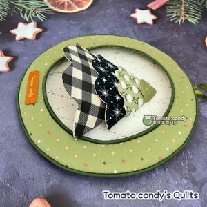 Pode incluir: Um ornamento de tecido verde com uma aplicação de árvore de tecido xadrez branco e preto. O ornamento tem um fundo branco com pequenas bolinhas verdes, laranja e azuis. O texto "Tomato candy" está escrito em coreano no ornamento.