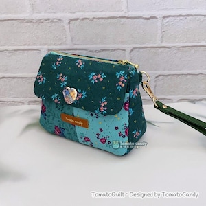 Può includere: Una borsa trapuntata con motivo floreale nei toni del verde acqua e verde. Ha una chiusura con patta, una cerniera dorata e un bottone a forma di cuore. È attaccato un cinturino da polso verde. Il testo "Tomato candy" è visibile su una piccola etichetta.