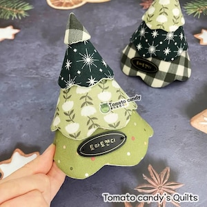 Könnte beinhalten: Handgefertigte Weihnachtsbaumdekorationen aus Stoff in Grün-, Weiß- und Schwarztönen. Die Bäume haben ein mehrschichtiges Design mit verschiedenen Mustern, darunter Blumen, Schneeflocken und Karos. Jeder Baum hat ein kleines, ovales Etikett mit Text. Der Text "Tomato candy's Quilts" ist ebenfalls sichtbar.