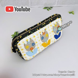 May include: A black and white quilted zippered pouch with a floral design. The pouch features three yellow, blue, and orange tulips with a white background. The pouch has a gold zipper and a black and white floral pattern on the back. The pouch is labelled "Tomato Candy" and has a link to the Etsy shop "tomatoquilt".