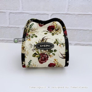 May include: A small, floral-patterned pouch with a black border. The pouch features a cream-colored background with a repeating pattern of red, white, and green flowers. The text "토마토캔디" is on a black oval label.