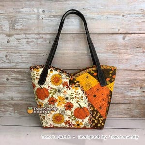 May include: A quilted tote bag featuring a patchwork design with autumn-themed fabric patterns including orange pumpkins, yellow flowers, and brown leaves on cream and orange backgrounds. The bag has two dark brown leather straps and a logo.