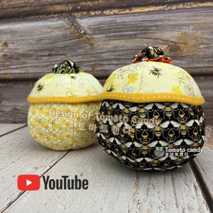 Peut inclure: Deux coussins d'épingle ronds en tissu avec des motifs d'abeilles. Un coussin d'épingle est jaune avec un motif de nid d'abeilles et l'autre est noir avec un motif d'abeilles. Les coussins d'épingle sont sur une surface en bois. Le texte "Design of Tomato Candy" est sur les coussins d'épingle.