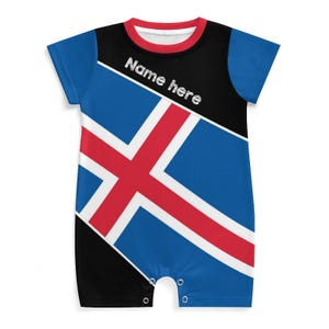 Peut inclure: Un body pour bébé bleu, noir et rouge avec le drapeau islandais. Le body a des manches courtes et un col rond bordé de rouge. Le texte "Name here" est imprimé sur le haut de la poitrine.